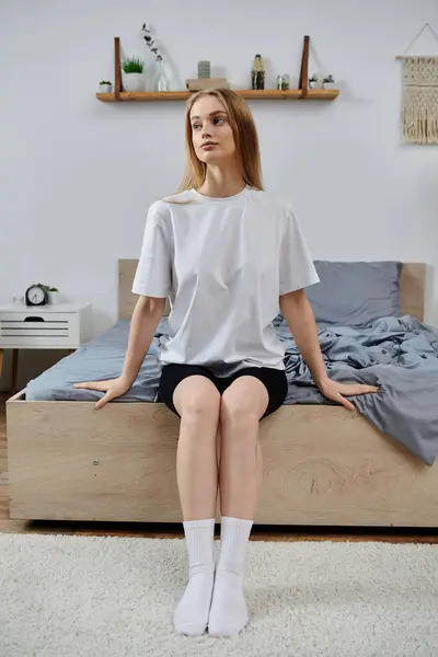 Young woman on bed in white t-shirt and black shorts looks to side. — Stock Photo