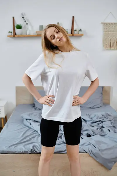 A young woman in a white t-shirt and black shorts stretches in her bedroom. — Stock Photo