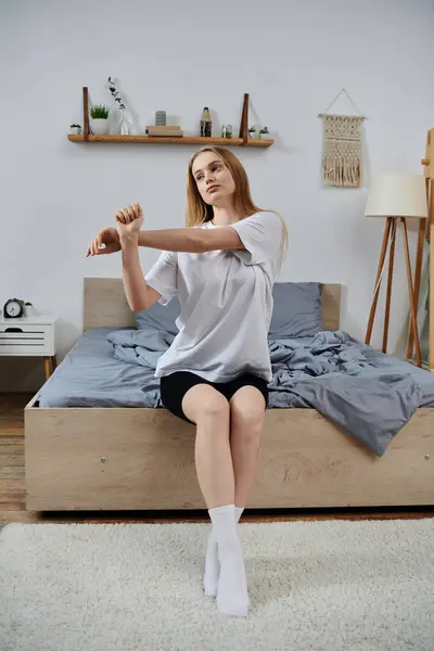 Una giovane donna in t-shirt bianca e pantaloncini neri si siede su un letto che si estende nella sua casa. — Foto stock