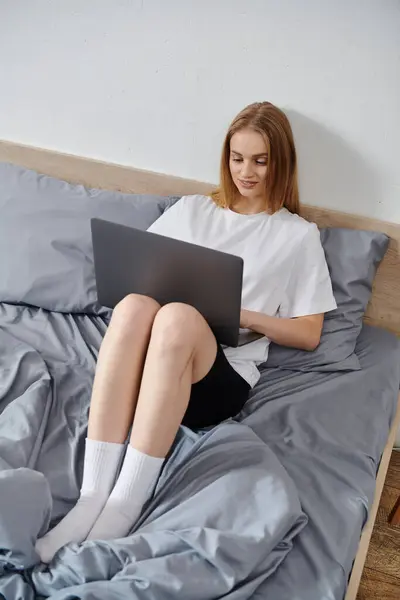 A young woman sits on her bed, working on a laptop. — Stock Photo