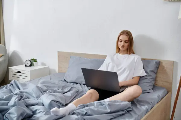 A young woman sits on a bed in a bedroom, using a laptop. — Stock Photo
