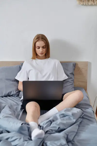 A young woman sits on a bed, using a laptop, surrounded by soft blankets. — Stock Photo