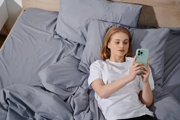 Una giovane donna giace a letto, scorrendo il suo telefono, godendo un momento di pace. — Foto stock