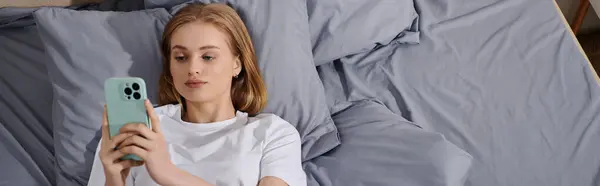 A young woman lies in bed, engrossed in her phone. — Stock Photo