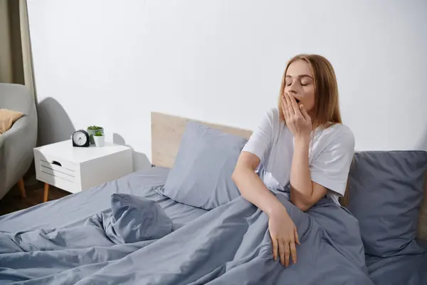 A young woman yawns while sitting on a bed in her bedroom. — Stock Photo