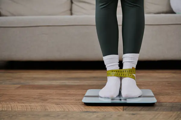 A young woman stands on a scale, a measuring tape tied around her ankles, in her home. — Stock Photo