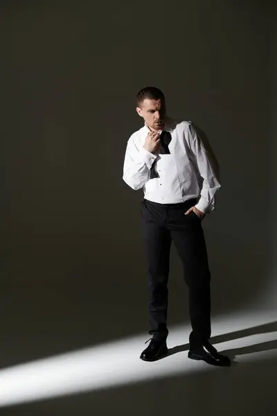 A man in a white shirt and black bow tie stands in a beam of light, adjusting his collar. — Stock Photo