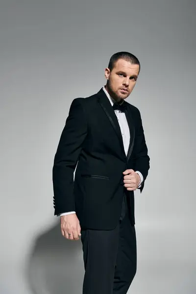 A man in a black tuxedo stands against a grey background, adjusting his jacket. — Stock Photo