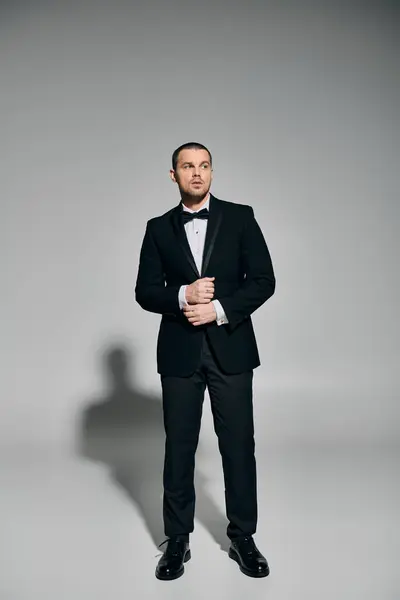 A man in a black tuxedo stands against a grey backdrop, looking up and adjusting his jacket. — Stock Photo