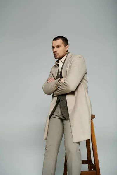 A stylish man in a beige coat stands with his arms crossed, leaning against a wooden stool. — Stock Photo