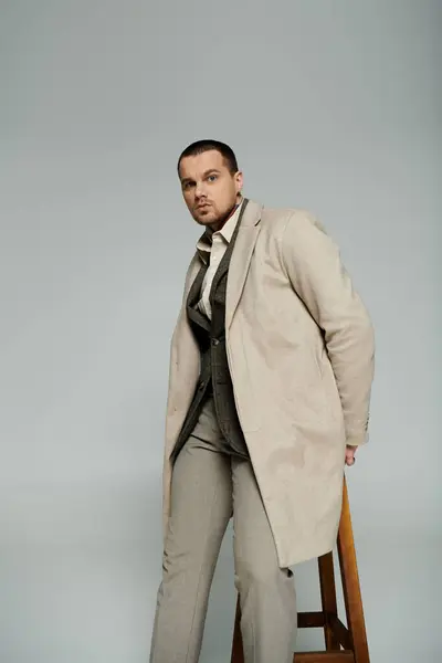 A stylish man in a beige coat leans casually on a wooden stool. — Stock Photo