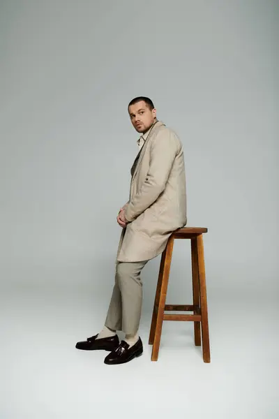 Stylish man in beige coat exudes confidence, sitting casually on stool. — Stock Photo