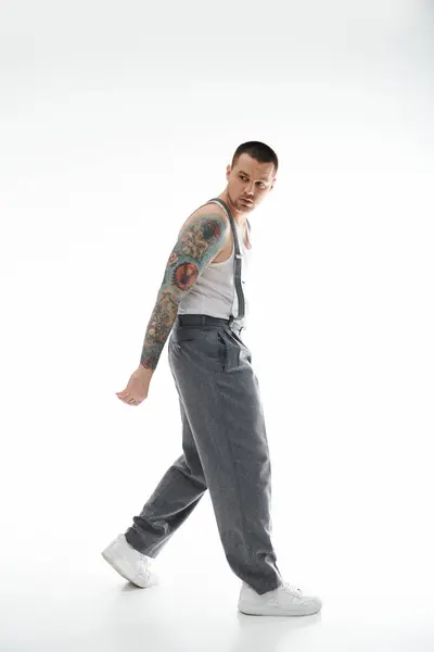 A man with a full sleeve tattoo walks confidently in a white tank top and grey slacks. — Stock Photo