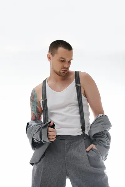 Un hombre con estilo en tirantes y una camiseta blanca se levanta con confianza, sosteniendo una chaqueta en una mano. — Stock Photo