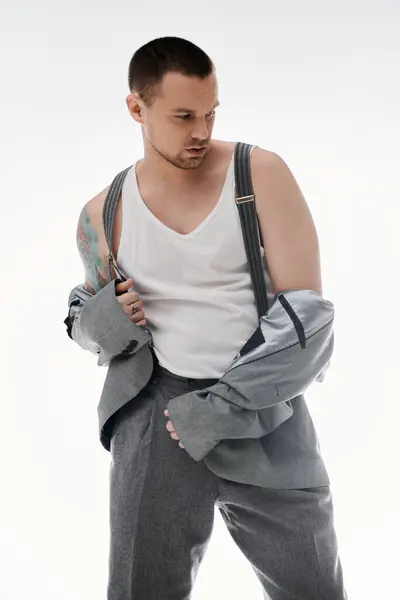 A man in a white tank top and suspenders adjusts his gray suit jacket against a white backdrop. — Stock Photo