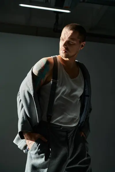 Man with buzz cut and tattoos in studio, wearing white tank top, suspenders, gray jacket. — Stock Photo