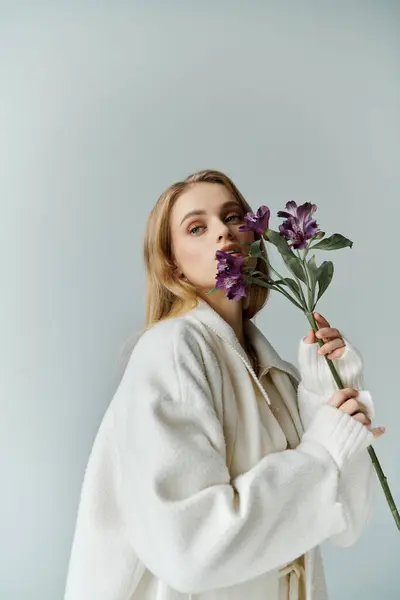Una mujer con un abrigo blanco sostiene una sola ramita de flores de color lila en su cara. - foto de stock