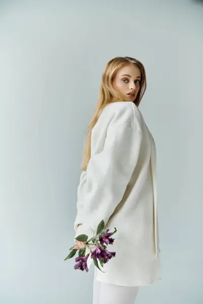Una mujer joven en un suéter blanco sostiene flores púrpuras detrás de su espalda, mirando por encima de su hombro. - foto de stock