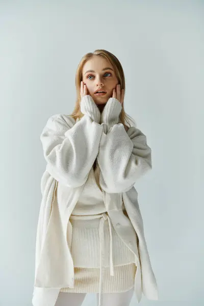 A young woman in a white knitted outfit stands with her hands on her cheeks, lost in thought. — Stock Photo