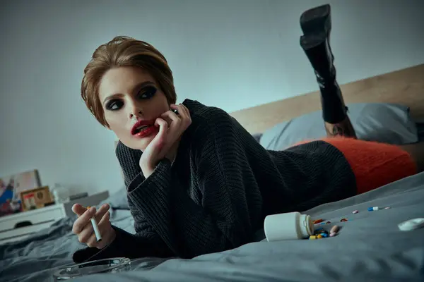 A woman with dark makeup and a stylish sweater lies on a bed, surrounded by scattered pills. — Stock Photo
