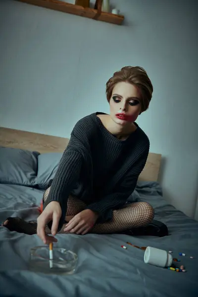 Uma jovem se senta em uma cama, com os olhos caídos, cercada por comprimidos espalhados e um cinzeiro. — Fotografia de Stock