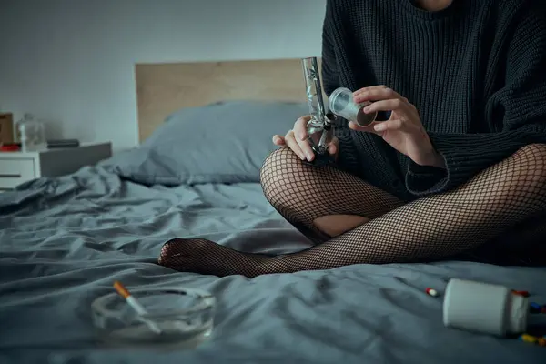 Женщина в черном свитере и чулках из рыбной сети сидит на кровати, держа бонг. — стоковое фото