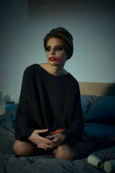 Grungy woman in dark sweater sits on bed, fishnet stockings contrasting soft bedding. — Stock Photo