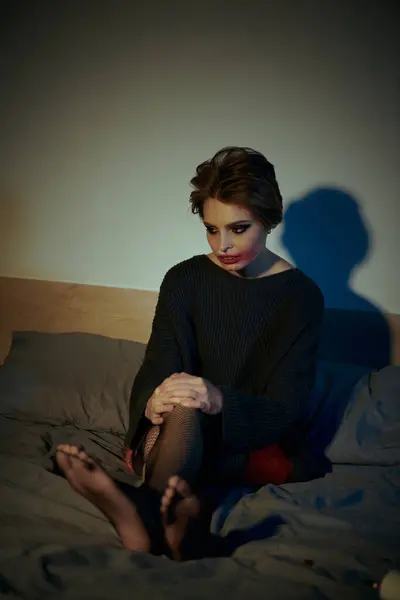 A woman with dark hair and striking makeup sits on a bed, her bare feet resting on the sheets. — Stock Photo