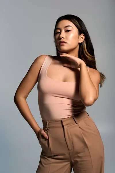 Asian woman wears pink tank top and brown pants, posing confidently against gray background. — Stock Photo