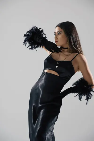 A young woman in a black satin dress and feather gloves poses against a neutral backdrop. — Stock Photo