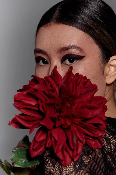 Asian woman in black lace dress, red flower over mouth, looks at viewer. — Stock Photo