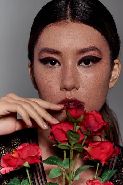 Une femme asiatique aux cheveux foncés et au maquillage audacieux sourit avec des roses rouges. — Photo de stock