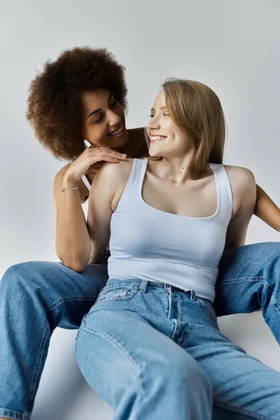 Dos mujeres en jeans y camisetas sin mangas se sientan sobre un fondo gris, sonriéndose una a la otra. - foto de stock