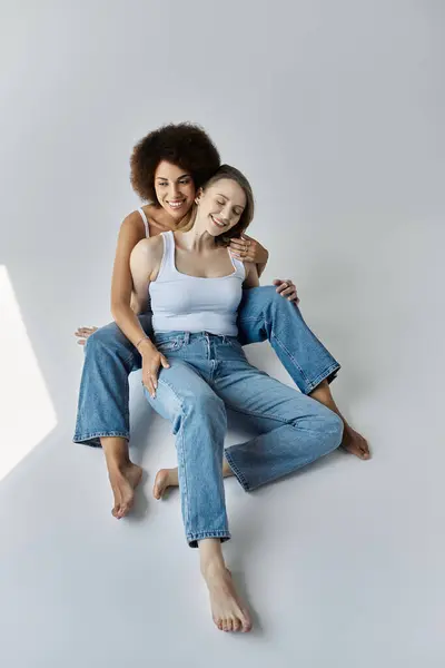 Um casal lésbico, usar tops tanque e jeans, rindo juntos enquanto deitado em um chão cinza. — Fotografia de Stock