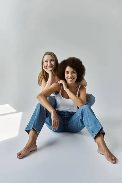 Deux femmes, l'une aux cheveux blonds et l'autre aux cheveux bruns bouclés, s'assoient sur un sol blanc en jeans et débardeurs blancs. — Photo de stock