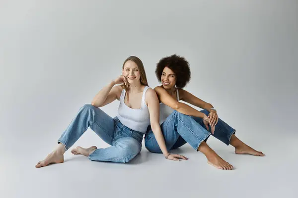 Due donne che indossano canottiere bianche e jeans, sedute su uno sfondo grigio. — Foto stock