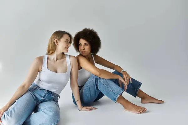 Two women, one with blonde hair and one with curly black hair, are sitting together in jeans and tank tops, looking at each other. — Stock Photo