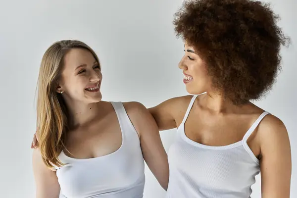 Two women wearing white tank tops smile at each other against a gray background. — Stock Photo