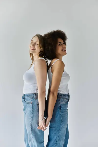 Two women, one Black and one white, stand back-to-back, smiling, wearing white tank tops and blue jeans. — Stock Photo