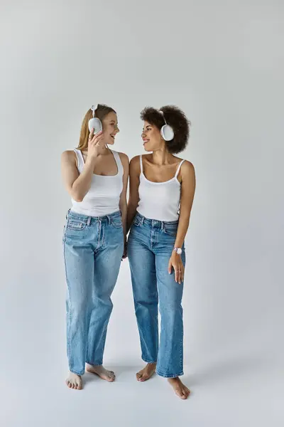 Two women wearing white tank tops and jeans smile at each other while wearing headphones. — Stock Photo