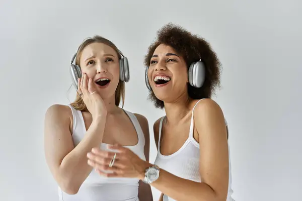 Due donne in canottiera bianca, una con i capelli biondi e una con i capelli castani ricci, ridono e cantano insieme alla musica indossando le cuffie. — Foto stock