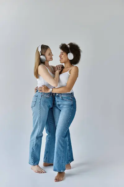 Two women in white tank tops and jeans embrace against a grey backdrop. — Stock Photo