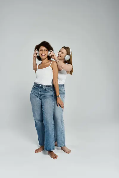 Two women, one Black and one White, stand together barefoot while wearing white tank tops and jeans, listening to music through headphones. — Stock Photo