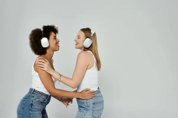 Due donne in canottiera bianca e jeans ridono indossando le cuffie. — Foto stock