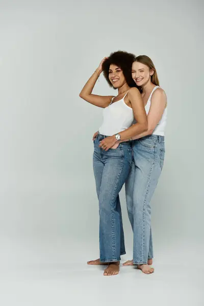 Duas mulheres, uma preta e uma branca, ficam descalças em jeans e tops de tanque, sorrindo e abraçando. — Fotografia de Stock