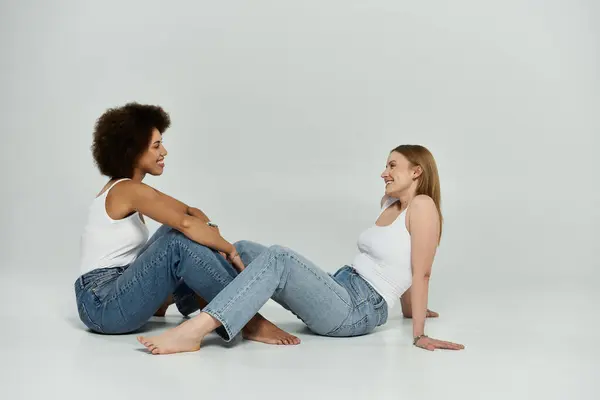 Two women in white tank tops and jeans laugh and look at each other on a white background. — Stock Photo