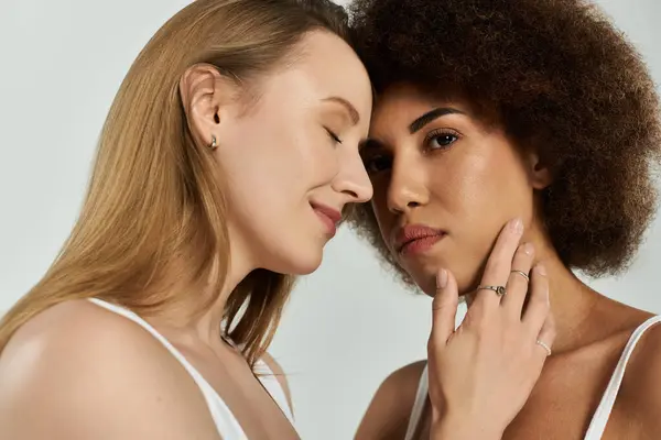 Two women in white tank tops and jeans embrace in a tender moment, their foreheads touching against a gray background. — Stock Photo