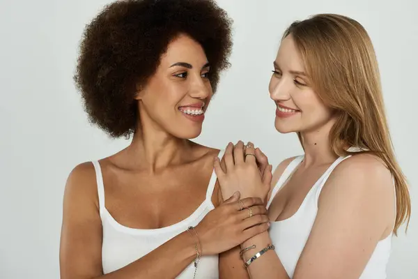 Two women in white tank tops and jeans, one with curly hair, look at each other lovingly. — Stock Photo