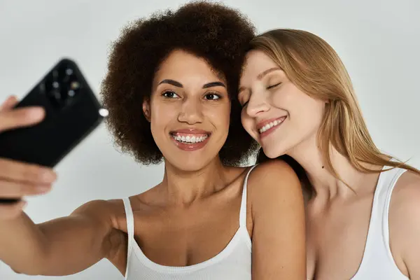 Due donne, una con i capelli ricci scuri e l'altra con lunghi capelli biondi, entrambe in canottiera bianca, si fanno un selfie. Sorridono e sembrano felici. — Foto stock
