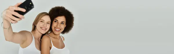 Deux femmes, une blanche et une noire, vêtues de débardeurs blancs et souriantes, prennent un selfie ensemble devant un fond gris. — Photo de stock